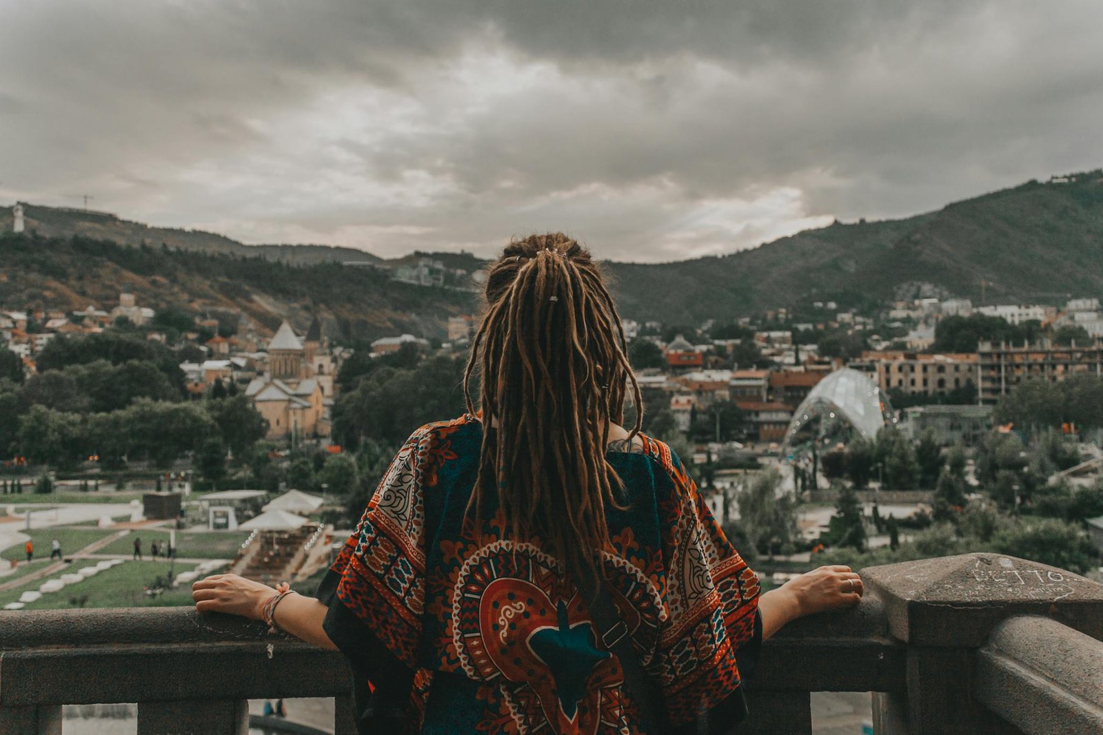 A person with dreadlocks overlooking the scenic cityscape of Tbilisi, Georgia, featuring mountains and architectural landmarks.